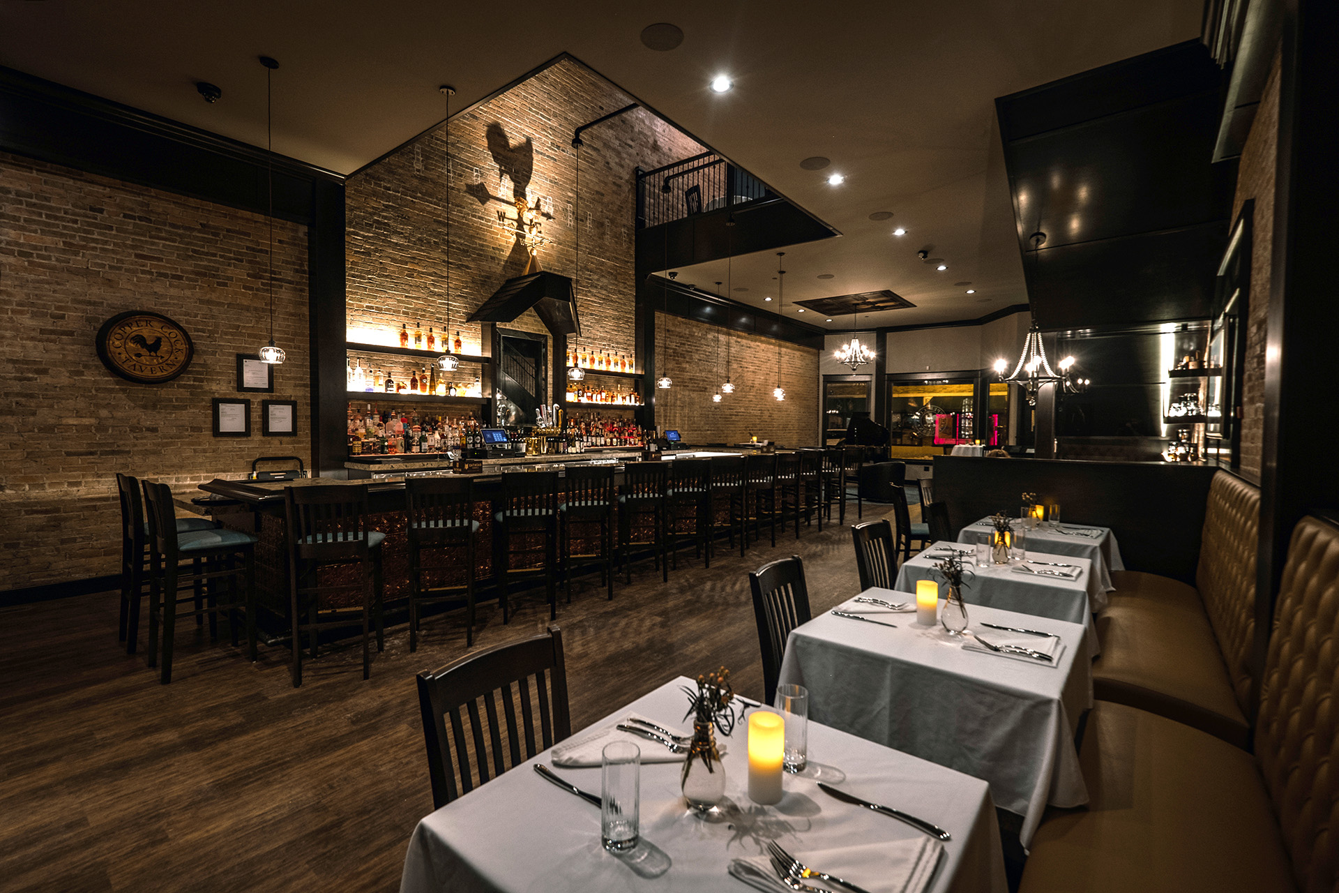 Copper Cock Tavern dining room and bar, featuring exposed brick, warm lighting, and the circular rooster wall mark